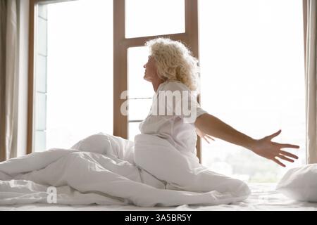 Eine erwachte, alte Hotelgast sitzt auf dem Bett und streckt die Arme Stockfoto