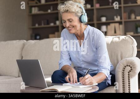 Ältere Frau mit Kopfhörern sehen Webinar über Notizen von Notizbüchern an Stockfoto
