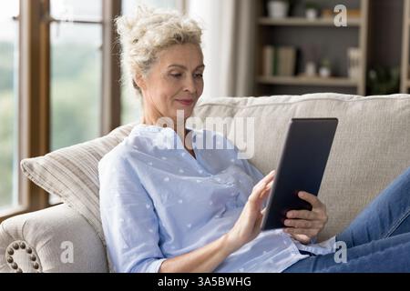Lächelnde, reife Dame, selbstbewusster Tablet-Benutzer, der sich auf einem gemütlichen Sofa ausruhen kann Stockfoto