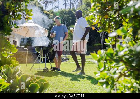 Ältere Freunde genießen Barbecue im Freien, halten Getränke und plaudern im sonnigen Hinterhof Stockfoto