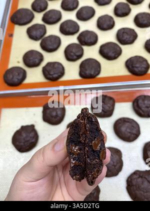 Nahaufnahme einer Hand, die einen Schokoladenkeks in zwei Hälften hält, und ein leckeres Schokoladeninnere enthüllt. Im Hintergrund sind mehrere ähnliche Cookies pla Stockfoto