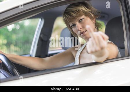 Attraktive blonde junge Frau, die ein Auto fährt und den Mittelfinger zeigt Stockfoto
