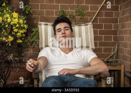Hübscher dunkelhaarigen jungen Mann trägt einen v-Ausschnitt T-shirt weiß beim Rauchen einer Zigarette in der einen Balkon auf Stuhl sitzend Stockfoto
