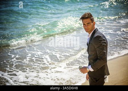 Schönen jungen Mann im klassischen Anzug am Strand Sonnenbrillen beim Blick in die Kamera halten. Wellen des Meeres im Hintergrund Stockfoto