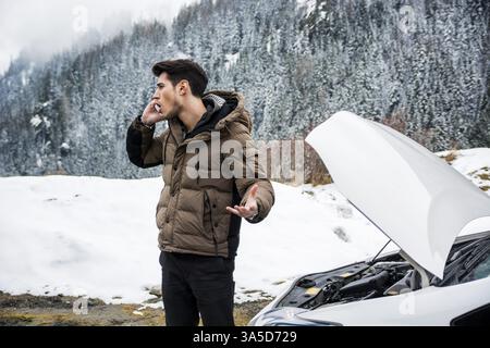 Junger Mann, der auf dem Handy in der Nähe eines Autos mit offener Haube spricht. Schneebedeckter Wald im Hintergrund Stockfoto
