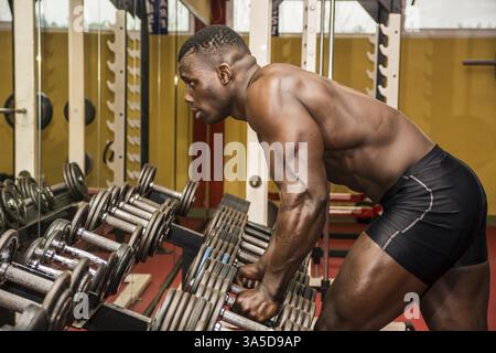 Schöner schwarzer Bodybuilder, der nach dem Training im Fitnessstudio ausgeruht und müde auf Gewichten gestützt ist Stockfoto