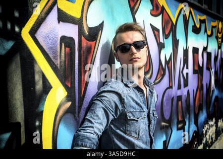 Hübscher blonder junger Mann an bunten Graffiti-Wand, Jeanshemd tragen Stockfoto