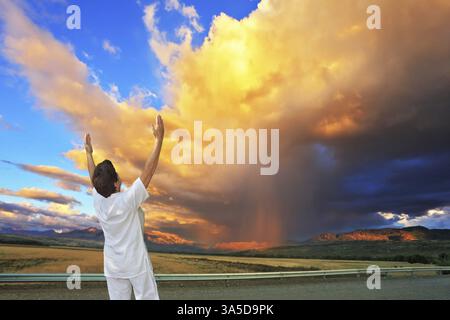Riesige Wolke, beleuchtet durch den Sonnenuntergang. Yoga auf der Straße. Ältere Frau in weiß macht Asana Sun Gruß auf der Autobahn Stockfoto
