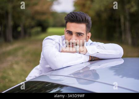 Porträt eines jungen, attraktiven Mannes im Business-Anzug, der sich auf seinem neuen, stilvollen Auto auf dem Land lehnt Stockfoto