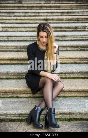 Porträt von attraktive blonde junge Frau im schwarzen Kleid, im freien Blick in die Kamera, auf großen alten Steintreppen Stockfoto