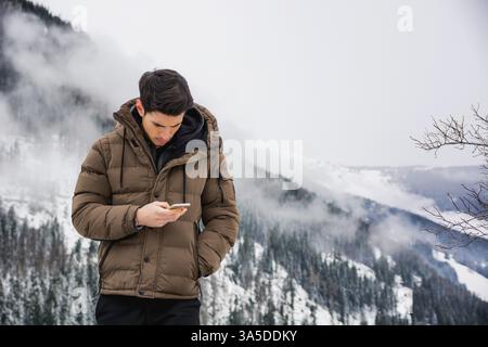 Dunkelhaariger, hübscher junger Mann in Winteroberbekleidung mit Handy oder Smartphone, draußen am Berg mit verschneiten Landschaften dahinter Stockfoto