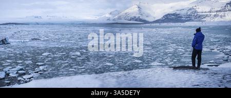 Ganzkörper-Rückblick auf nicht wiedererkennbaren männlichen Touristen in Oberbekleidung, die in der Nähe des eisigen Meeres stehen und die malerische Landschaft der schneebedeckten Berge in Island bewundern Stockfoto