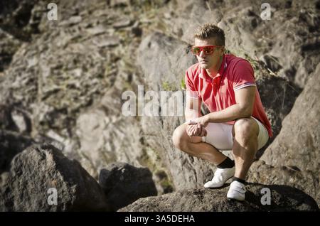 Hübscher junger Mann sitzt auf Felsen mit Sonnenbrille, Blick auf einer Seite Stockfoto