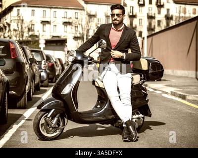 Junger, stilvoller Mann im Anzug und Sonnenbrille, der neben seinem klassischen italienischen Roller auf einer leeren Straße in der sonnigen Stadt steht Stockfoto