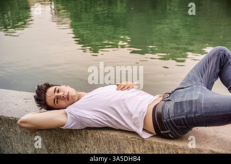 Hübscher junger Mann an einem See', am Ufer oder am Flussufer an einem sonnigen, friedlichen Tag, liegend Stockfoto