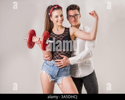 Junger gutaussehender Mann in Hemd und Brille, der ein junges trendiges Model umschließt, mit Hantel, die Bizeps zeigt Stockfoto