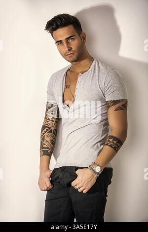 Stattliche tätowierten jungen Mann mit grauen T-Shirt, stehend gegen den hellen Hintergrund im Studio shot Stockfoto