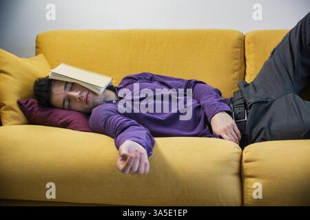 Überarbeiteter, müder junger Mann zu Hause schläft statt zu arbeiten oder zu studieren, ruht mit Kopf bedeckt mit offenem Buch Stockfoto