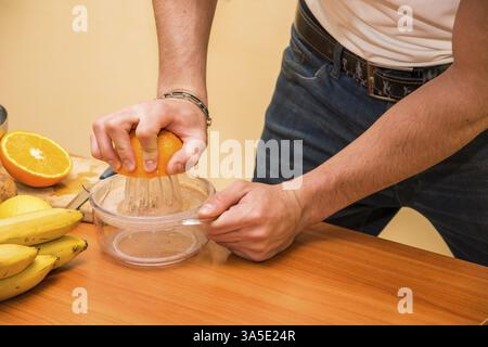 Junger Mann, der eine Orange quetscht, um gesunden Obstsalat oder Smoothie auf einer Küchentheke mit einer Auswahl an frischem Obst zuzubereiten Stockfoto