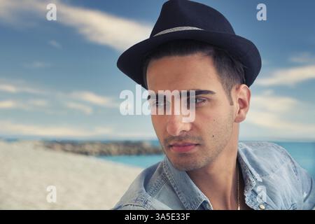 Hübscher junger Mann mit Jeansshirt und Fedorahut, der in Pose am Sandstrand sitzt und wegblickt Stockfoto