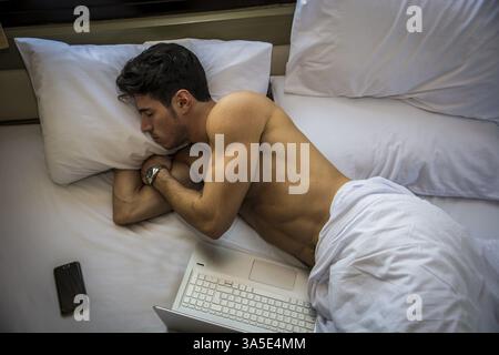 Der junge Mann mit dem dunklen Haar Schlaf im Bett neben Laptop und Handy, müde von zu viel Arbeit Stockfoto