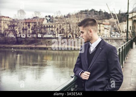 Dreiviertel langer, nachdenklicher, hellbrauner junger Mann mit grauer Jacke und Denim-Jeans, der am malerischen Fluss in Turin, Ita, steht Stockfoto