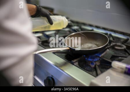 Der professionelle Koch trägt schwarze Handschuhe, gießt Öl aus einer Plastikflasche in eine Pfanne auf einem Gasherd und bereitet sich auf indische Gerichte in einem Restaurantset vor Stockfoto