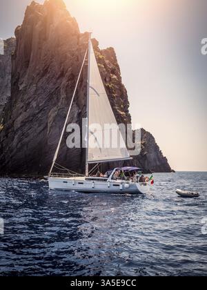 Moderne weiße Yacht segelt langsam im plätschernden Meer in der Nähe von rauem Felsen an wolkenlosem Abend im Resort Stockfoto