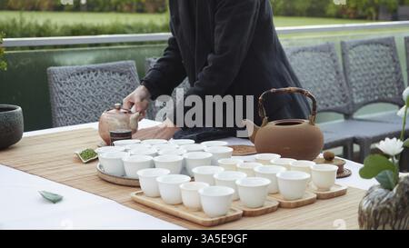 Eine zugeschnittene Person serviert Teeservice an einem Tisch im Freien. Terrakotta-Teekanne, weiße Tassen auf Holztabletts auf Bambusmatte Stockfoto