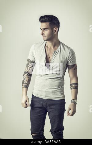 Stattliche tätowierten jungen Mann mit grauen T-Shirt, stehend gegen den hellen Hintergrund im Studio shot Stockfoto