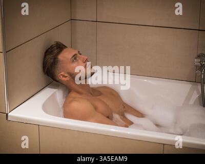 Ein hübscher junger Mann in der Badewanne zu Hause, der badet und sich auf den Rand der Badewanne lehnt Stockfoto