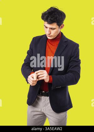 Hübscher junger Mann im Studio, der seine Jacke anpasst, den Ärmel mit ernstem Ausdruck ansieht, einen Rollkragenpullover auf gelbem Hintergrund trägt Stockfoto