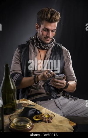 Modischer Mann als Pirate gekleidet, mit Handy mit Lederweste und Schal, der im Stuhl neben dem Tisch sitzt, mit leerer Flasche und verschiedenen Gegenständen Stockfoto