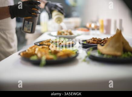 Der Koch mit schwarzen Handschuhen gießt eine Soße aus einer Flasche auf mehrere Teller indischer Speisen, darunter Samosas und Pakoras, die auf einem weißen Tisch angeordnet sind Stockfoto