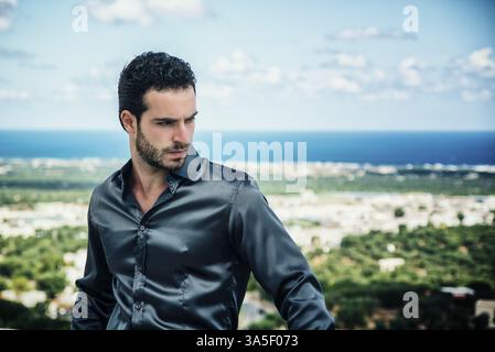 Porträt von eleganten attraktiver junger Mann mit lockigem Haar vor der Küste stehen und blickte mit ernstem Gesicht Stockfoto