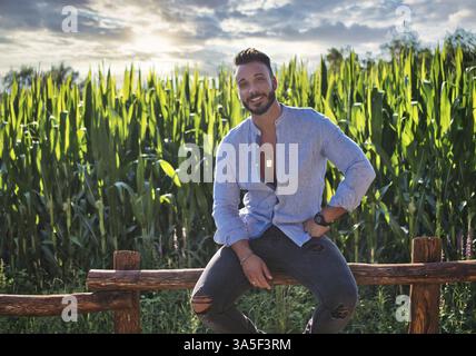 Hübscher Hunky junger Mann im Freien auf dem Land auf dem Holzzaun sitzend, während er die Kamera anschaut Stockfoto