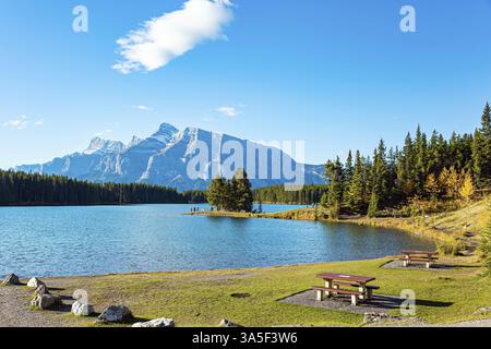 Gemütlicher Picknicktisch und Bänke aus Holz. Zwei Jack Lake. Der riesige Gletschersee reflektiert Sonne und Wolken. Reisen Sie nach Kanada, den Rocky Mountains Stockfoto