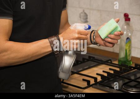 Nahansicht des jungen Mannes', s Arme Reinigungsfläche der Herdplatte in der Küche mit Schwamm - verantwortlicher Mann, der Hausarbeit in der ersten Wohnung macht Stockfoto