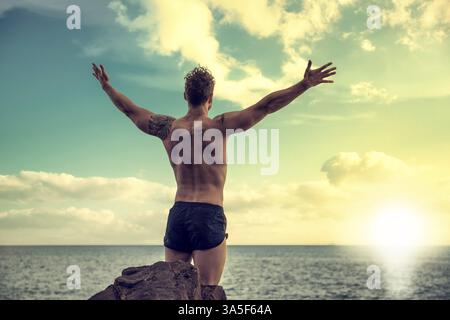 Muskulöser junger Mann am Strand, von hinten gesehen, mit offenen Armen, das Gefühl der Freiheit vor aufgehender oder untergehender Sonne genießt Stockfoto