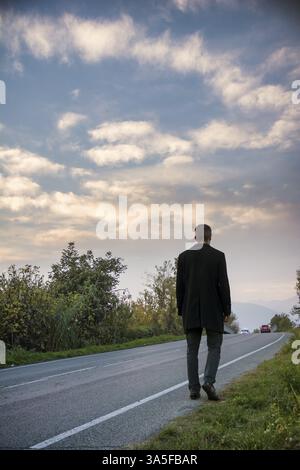 Rückansicht eines Mannes im Anzug, der auf einer Straße in ländlicher Umgebung läuft. Vertikale Außenaufnahme Stockfoto