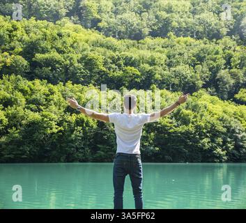 Junger Mann im T-Shirt, im Freien mit offenen Armen, der Freiheit vor dem See genießt, von hinten gesehen Stockfoto