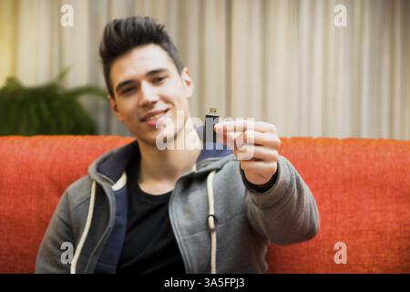 Hübscher lächelnder junger Mann zu Hause zeigt USB-Schlüssel in der Hand, sitzt auf einer couch Stockfoto