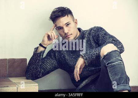 Hübscher weißer asiatischer junger Mann mit grauem Pullover und zerrissener Jeans, der auf hellem Hintergrund vor der Wand im Studio sitzt Stockfoto