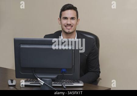 Freundlich, lächelnd jungen Geschäftsmann am Schreibtisch in seinem Büro mit Computer arbeiten Stockfoto
