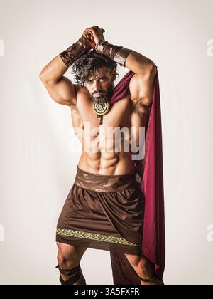 Schöner muskulöser Mann, der in römischem oder spartanischem Gladiatorenkostüm mit Schild und Schwert posiert, isoliert auf schwarzem Hintergrund im Studio Stockfoto
