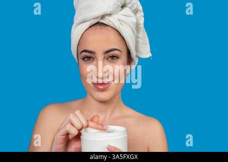 Schöne Frau, die ein Handtuch auf dem Kopf trägt und nach dem Duschen Feuchtigkeitscreme auf ihr Gesicht aufträgt Stockfoto