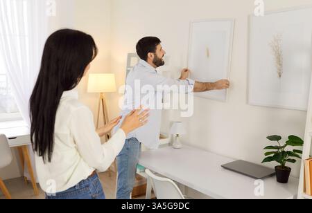 Fröhliches junges Paar, das ihr neues Zuhause zusammen dekoriert, ein Bild an die Wand hängt, als Team arbeitet. Stockfoto
