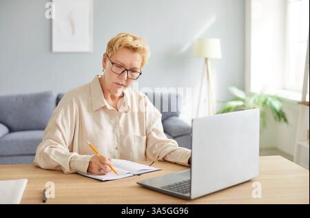 Ältere, beschäftigte, reife Frau, die eine Brille trägt, Notizen schreibt, einen Laptop benutzt, am Tisch sitzt Stockfoto
