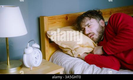 Junger Mann, der sich darum bemüht, aufzuwachen – schaut auf dem Wecker Schlummerlicht Stockfoto