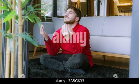 Junger Mann, der zu Hause betet, im Kreuz auf dem Boden sitzt, mit Händen zusammen in einer spirituellen und hingebungsvollen Geste Stockfoto